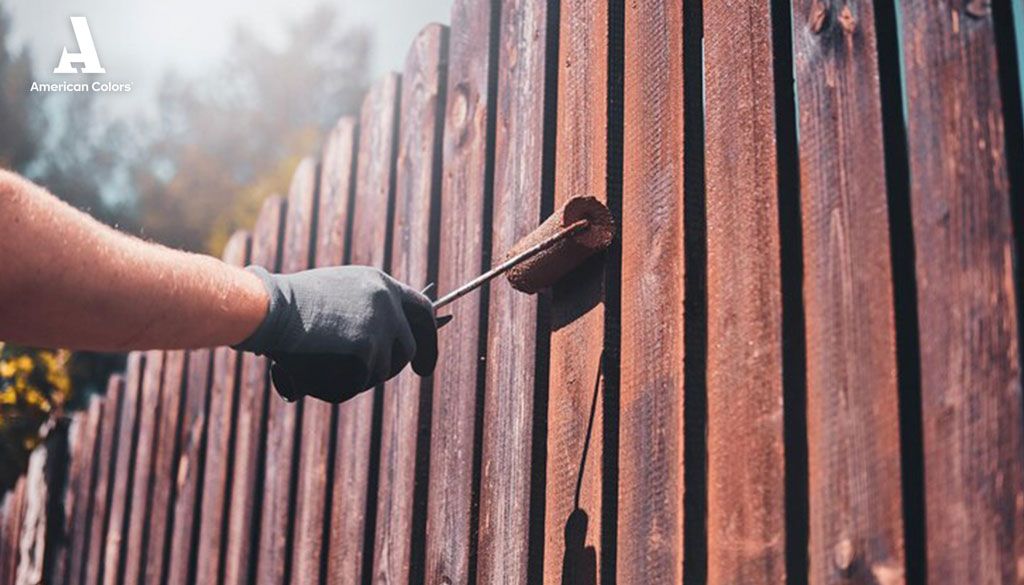 Mano con guante aplicando pintura protectora a una cerca de madera con un rodillo, destacando la importancia de pintura para exteriores.